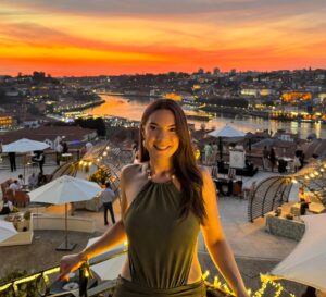 Headshot of Ashley Miller Daniels smiling in a green dress with a background of Porto.