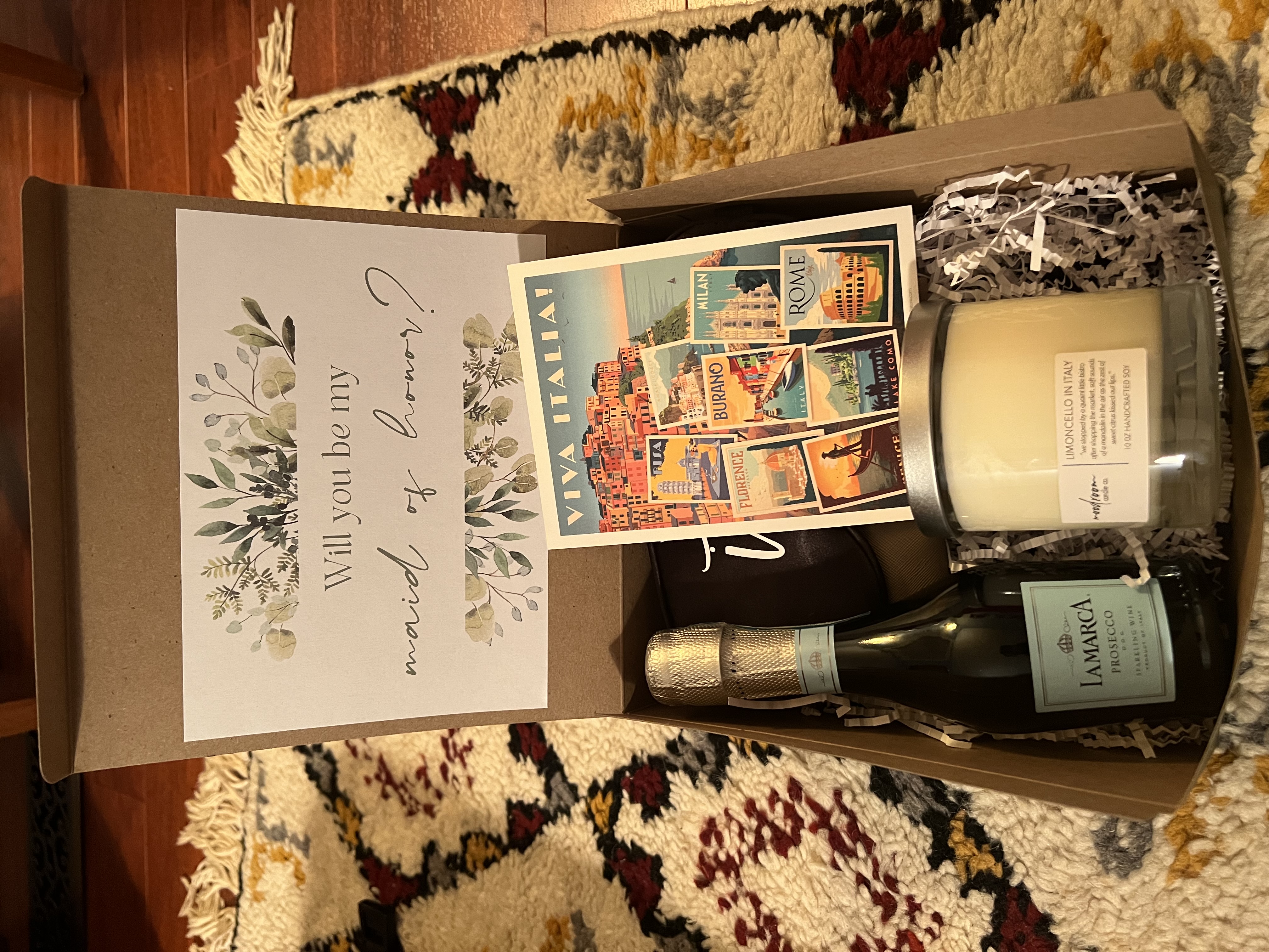 The interior of a curated bridesmaid proposal box featuring a limoncello candle, custom passport cover, and silk eye mask.