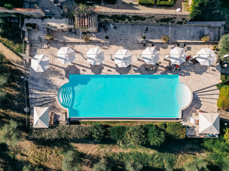 Aerial view of a large swimming pool with colorful umbrellas around it. The pool is surrounded by trees and lounge chairs. This is a wedding venue in italy, for a destination wedding in italy. wedding Venue in italy